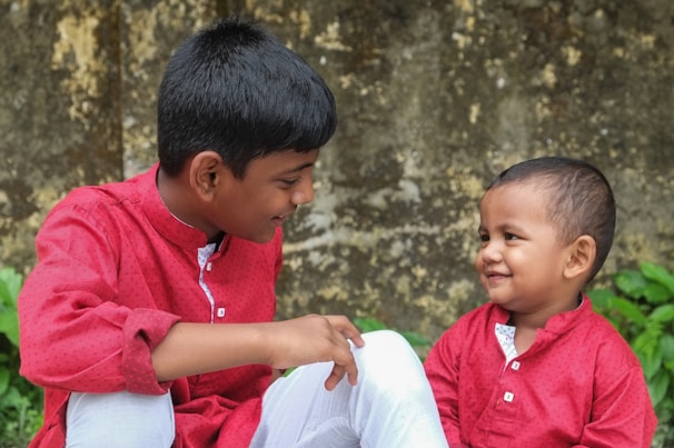 Two young children dressed in matching red shirts sit outdoors, sharing a moment of connection and joy. The older child is gazing at the younger with a smile, while the younger child looks back with a playful expression. A weathered wall and some greenery are visible in the background.