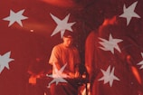 A musician is performing under dim red lighting, playing a keyboard. White star shapes overlay the scene, creating a dreamy, artistic effect. The musician is focused on playing, surrounded by other band members and equipment.
