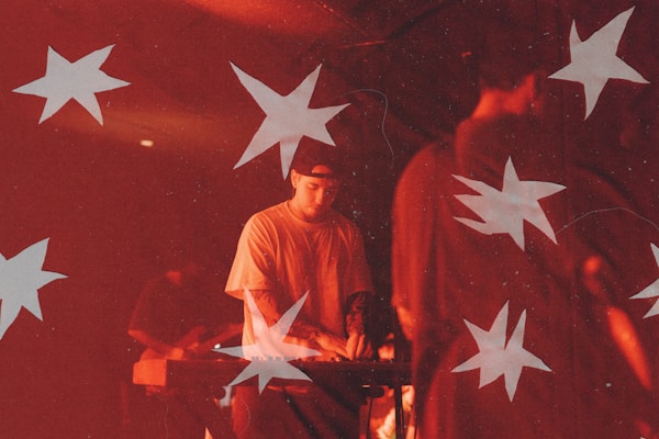 A musician is performing under dim red lighting, playing a keyboard. White star shapes overlay the scene, creating a dreamy, artistic effect. The musician is focused on playing, surrounded by other band members and equipment.