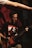 A man wearing glasses and a Metallica shirt is playing a guitar. His focus seems to be on the instrument as he stands against a dimly lit background with red and black tones. Another person's arm extends into the frame from the top left corner.