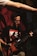 A man wearing glasses and a Metallica shirt is playing a guitar. His focus seems to be on the instrument as he stands against a dimly lit background with red and black tones. Another person's arm extends into the frame from the top left corner.