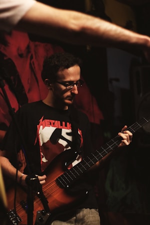 A man wearing glasses and a Metallica shirt is playing a guitar. His focus seems to be on the instrument as he stands against a dimly lit background with red and black tones. Another person's arm extends into the frame from the top left corner.