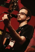 A musician with glasses plays a guitar and sings into a microphone. The background is a warmly lit stage with red and dark tones, subtle lighting highlights the musician's face and instrument.