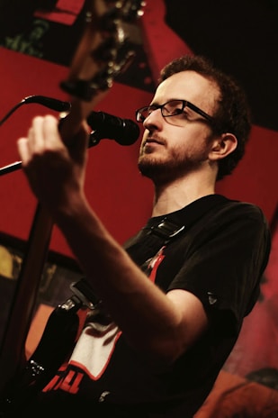 A musician with glasses plays a guitar and sings into a microphone. The background is a warmly lit stage with red and dark tones, subtle lighting highlights the musician's face and instrument.