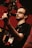 A musician with glasses plays a guitar and sings into a microphone. The background is a warmly lit stage with red and dark tones, subtle lighting highlights the musician's face and instrument.