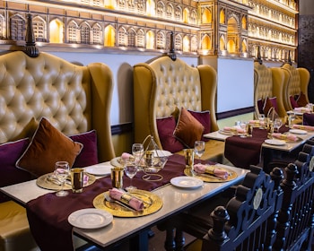 An elegantly set dining table in a restaurant with plush leather chairs. The chairs are high-backed and tufted in a mustard color, complemented by burgundy and dark brown cushions. The tables are adorned with gold decorative plates, gold utensils, and neatly rolled napkins. A rich burgundy table runner adds a touch of sophistication. The wall features intricate architectural decor resembling palatial windows with warm ambient lighting.