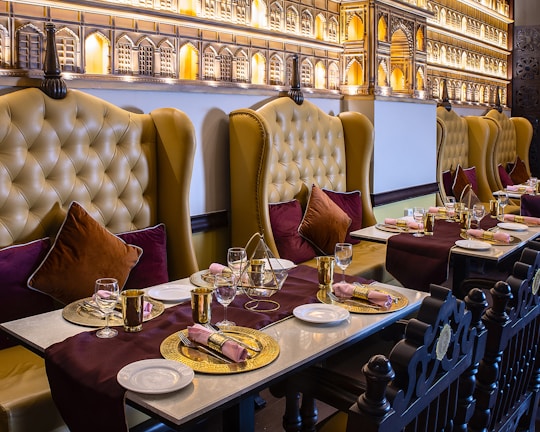An elegantly set dining table in a restaurant with plush leather chairs. The chairs are high-backed and tufted in a mustard color, complemented by burgundy and dark brown cushions. The tables are adorned with gold decorative plates, gold utensils, and neatly rolled napkins. A rich burgundy table runner adds a touch of sophistication. The wall features intricate architectural decor resembling palatial windows with warm ambient lighting.
