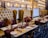 An elegantly set dining table in a restaurant with plush leather chairs. The chairs are high-backed and tufted in a mustard color, complemented by burgundy and dark brown cushions. The tables are adorned with gold decorative plates, gold utensils, and neatly rolled napkins. A rich burgundy table runner adds a touch of sophistication. The wall features intricate architectural decor resembling palatial windows with warm ambient lighting.
