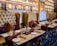 An elegantly set dining table in a restaurant with plush leather chairs. The chairs are high-backed and tufted in a mustard color, complemented by burgundy and dark brown cushions. The tables are adorned with gold decorative plates, gold utensils, and neatly rolled napkins. A rich burgundy table runner adds a touch of sophistication. The wall features intricate architectural decor resembling palatial windows with warm ambient lighting.