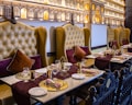 An elegantly set dining table in a restaurant with plush leather chairs. The chairs are high-backed and tufted in a mustard color, complemented by burgundy and dark brown cushions. The tables are adorned with gold decorative plates, gold utensils, and neatly rolled napkins. A rich burgundy table runner adds a touch of sophistication. The wall features intricate architectural decor resembling palatial windows with warm ambient lighting.