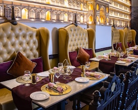 An elegantly set dining table in a restaurant with plush leather chairs. The chairs are high-backed and tufted in a mustard color, complemented by burgundy and dark brown cushions. The tables are adorned with gold decorative plates, gold utensils, and neatly rolled napkins. A rich burgundy table runner adds a touch of sophistication. The wall features intricate architectural decor resembling palatial windows with warm ambient lighting.