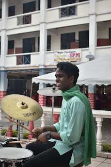 A person is smiling while playing drums, wearing a light green shirt and a matching scarf. The background features a multi-story building with open windows and a banner, as well as some red chairs under a canopy.