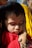 A young child with closed eyes rests their head on an adult's hand, which gently cradles the child's cheek. The child is wearing a red garment and has short, dark hair. Sunlight highlights the child's delicate facial features.