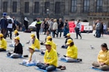 A group of people, mostly wearing yellow clothing and hats, are seated on mats practicing meditation in an outdoor public square. Nearby, other pedestrians walk by, and there are banners in the background with text related to stopping executions in Iran. The setting includes pigeons on the ground, and a police officer is visible among the crowd.