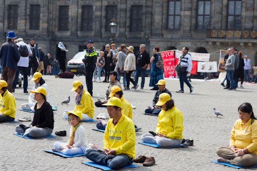 A group of people, mostly wearing yellow clothing and hats, are seated on mats practicing meditation in an outdoor public square. Nearby, other pedestrians walk by, and there are banners in the background with text related to stopping executions in Iran. The setting includes pigeons on the ground, and a police officer is visible among the crowd.