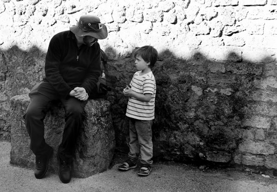 A person wearing a hat sits on a stone bench in a thoughtful pose, while a young child stands next to them, looking up. The background consists of a textured stone wall, casting shadows.