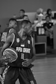 A basketball player wearing a jersey with the text 'Papua Barat' dribbles a basketball while focused on the game. The scene is monochrome, showing intensity and movement. Other players and spectators are visible in the background, watching the action.