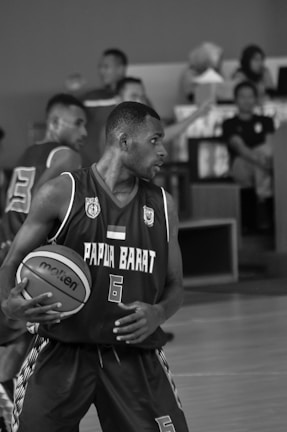 A basketball player wearing a jersey with the text 'Papua Barat' dribbles a basketball while focused on the game. The scene is monochrome, showing intensity and movement. Other players and spectators are visible in the background, watching the action.