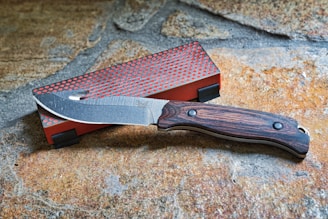 A knife with a wooden handle and a partially serrated stainless steel blade rests on a stone surface. Next to it is a rectangular red sharpening stone with a textured gray surface.