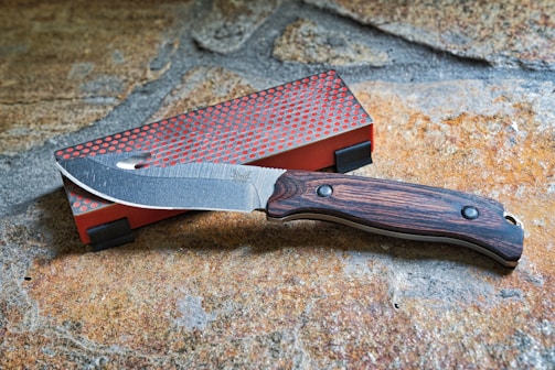 A knife with a wooden handle and a partially serrated stainless steel blade rests on a stone surface. Next to it is a rectangular red sharpening stone with a textured gray surface.