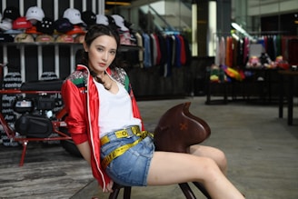 A woman is sitting on a wooden object shaped like an animal in a shop. She is wearing a red jacket with shiny details, a white shirt, and denim shorts with a yellow belt. Behind her, there is a display of various hats and a rack of clothing.