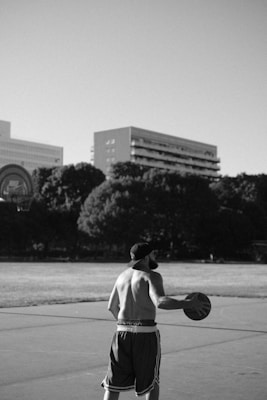 A shirtless man stands on a basketball court holding a basketball under one arm. He is looking towards the basketball hoop, which is visible in the background. The setting is outdoors with trees and large buildings in the distance. The image is in black and white, giving it a classic and timeless feel.