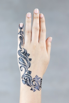 A hand with intricately designed henna or temporary tattoo art on the back of it, featuring elaborate black patterns with curving lines and geometric shapes. The skin tone is light, and the nails are unpainted and clean. The background is a plain gray, providing contrast to the detailed design.