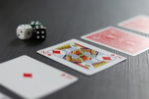 A King of Diamonds playing card is prominently displayed in the foreground on a dark surface, with two red-backed cards beside it. Two dice, one white and one black, are positioned to the left.
