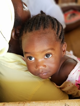 A young child with braided hair is resting on an adult's shoulder. The child has bright eyes and is wearing a garment with small pink hearts. The adult is wearing a light yellow garment, and their face is partially visible. The background is softly blurred, focusing on the child.