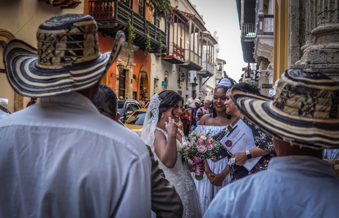 A bride in a white lace wedding dress with a bouquet of pink roses surrounded by guests wearing traditional Colombian sombrero vueltiao hats on a colourful Cartagena Old City street