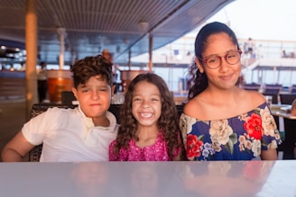 Three children sit together at a table in a well-lit area. The child on the left wears a white shirt and has a neutral expression. The middle child, in a glittery pink top, smiles brightly. The child on the right has glasses and wears a floral-patterned off-the-shoulder dress, smiling softly. The background shows a decked area with railings and people blurred in the distance.