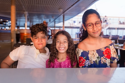 Three children sit together at a table in a well-lit area. The child on the left wears a white shirt and has a neutral expression. The middle child, in a glittery pink top, smiles brightly. The child on the right has glasses and wears a floral-patterned off-the-shoulder dress, smiling softly. The background shows a decked area with railings and people blurred in the distance.