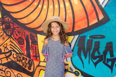 A smiling young girl in a colorful, comfortable dress standing against a vibrant mural.