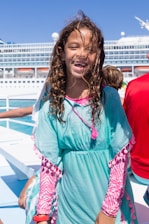 A young girl with long, curly hair smiles broadly. She wears a light teal dress with pink accents and a pink zebra-patterned shirt underneath. Behind her is a large white cruise ship set against a clear blue sky.