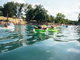 People are enjoying a sunny day at a natural swimming spot, floating on inflatable tubes. The area is surrounded by lush trees and a grassy hill where more people are relaxing and socializing.