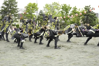 A group of people dressed in coordinated costumes with swirling yellow patterns performing an energetic dance on an outdoor open space. Lush green trees and traditional-style houses are in the background, with red and white lanterns strung across the area.