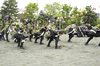 A group of people dressed in coordinated costumes with swirling yellow patterns performing an energetic dance on an outdoor open space. Lush green trees and traditional-style houses are in the background, with red and white lanterns strung across the area.