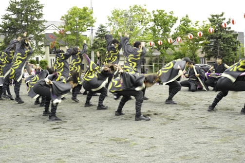 A group of people dressed in coordinated costumes with swirling yellow patterns performing an energetic dance on an outdoor open space. Lush green trees and traditional-style houses are in the background, with red and white lanterns strung across the area.
