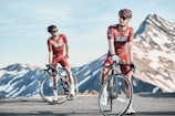 Two cyclists in red biking gear are positioned on a mountain road with snow-capped peaks in the background. Both are wearing helmets and sunglasses, standing next to their road bikes. The sky is clear, and the scene conveys a sense of freedom and adventure.