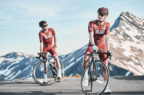 Two cyclists in red biking gear are positioned on a mountain road with snow-capped peaks in the background. Both are wearing helmets and sunglasses, standing next to their road bikes. The sky is clear, and the scene conveys a sense of freedom and adventure.