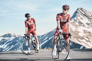 Two cyclists in red biking gear are positioned on a mountain road with snow-capped peaks in the background. Both are wearing helmets and sunglasses, standing next to their road bikes. The sky is clear, and the scene conveys a sense of freedom and adventure.
