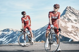 Two cyclists in red biking gear are positioned on a mountain road with snow-capped peaks in the background. Both are wearing helmets and sunglasses, standing next to their road bikes. The sky is clear, and the scene conveys a sense of freedom and adventure.