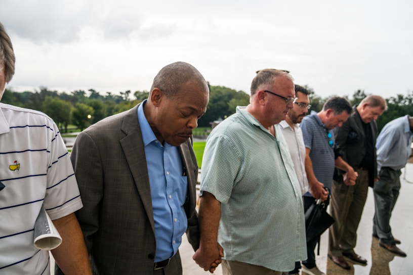 A group of men standing outdoors, holding hands with heads bowed, suggesting a moment of silence or prayer. They are dressed in casual and business attire, with a green landscape visible in the background, under an overcast sky.