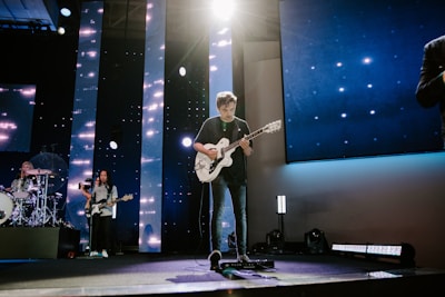A band performs on a stage lit with dynamic lighting. The guitarist is at the center, standing under a bright spotlight while playing an electric guitar. To the left, the drummer is visible behind a drum kit, and another musician plays a bass guitar. The stage has vertical LED lights and a screen backdrop displaying star-like visuals.
