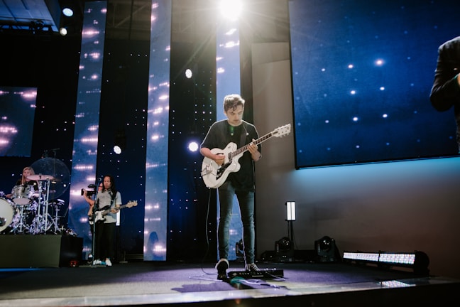 A band performs on a stage lit with dynamic lighting. The guitarist is at the center, standing under a bright spotlight while playing an electric guitar. To the left, the drummer is visible behind a drum kit, and another musician plays a bass guitar. The stage has vertical LED lights and a screen backdrop displaying star-like visuals.