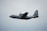A military cargo aircraft, specifically a U.S. Air Force plane, is flying through a cloudy sky. The aircraft features a gray exterior with markings that include an American flag and the designation 'PR'.