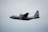 A military cargo aircraft, specifically a U.S. Air Force plane, is flying through a cloudy sky. The aircraft features a gray exterior with markings that include an American flag and the designation 'PR'.