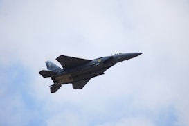 A military jet fighter is soaring through a cloudy sky. The aircraft is angled upward, displaying its streamlined design and sharp wings. It appears to be an advanced tactical aircraft, painted in dark grey with some markings visible on the tail.