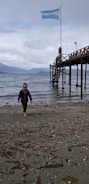 A child in a winter jacket and hat walks along a pebbly beach. A wooden pier extends over the water with an Argentine flag waving at its end. The background reveals a cloudy sky and distant mountains across the water.