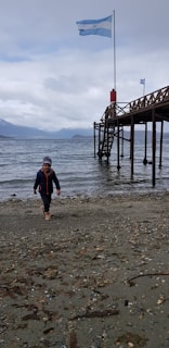 A child in a winter jacket and hat walks along a pebbly beach. A wooden pier extends over the water with an Argentine flag waving at its end. The background reveals a cloudy sky and distant mountains across the water.